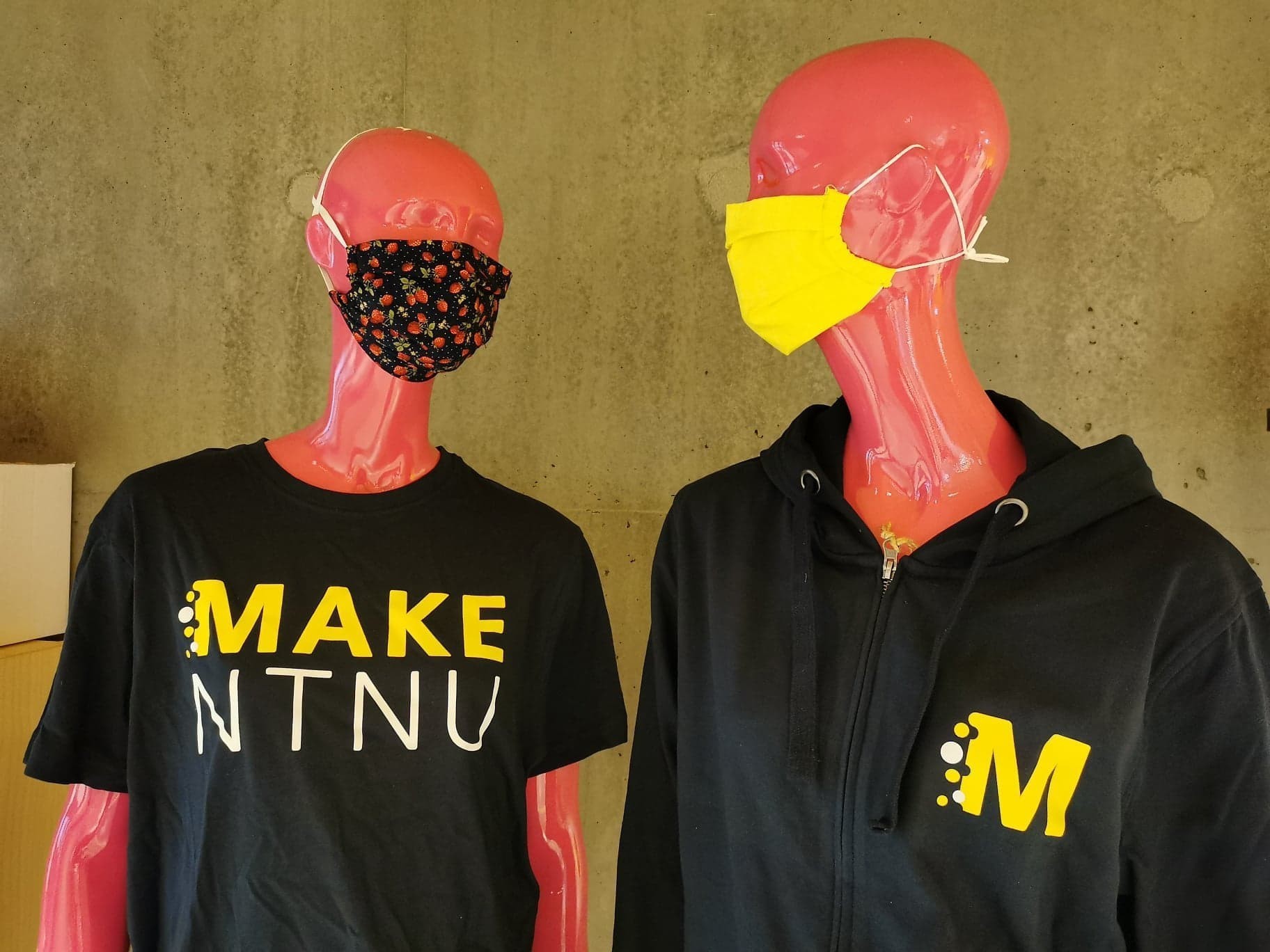Two pink mannequins with MAKE NTNU apparel; the first one is wearing a black face mask with a strawberry pattern, and the second one is wearing a yellow face mask